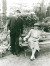 An elderly man stands with a cane next to a smiling woman on a garden bench, both dressed in suits and hats among green plants.