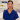 A female healthcare professional in blue scrubs smiles calmly in a well-equipped, clean clinical environment.