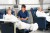 A nurse in blue scrubs speaks with an older woman in a reclining hospital chair; both patients rest comfortably with white blankets.