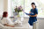 A nurse in blue scrubs smiles and records notes at a bedside where a red-haired woman sits up, with flowers and equipment nearby.