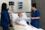 Two clinicians in blue scrubs support an older woman in a hospital bed, monitoring vital signs and communicating with care.