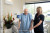 An elderly man using a walker receives attentive support from a smiling caregiver in a bright, welcoming hospital room.