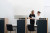 Two staff members review documents behind a reception counter in a clean, modern office. Empty chairs and workstations are visible.