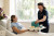 A hospital patient smiles at a staff member delivering a meal tray. Sunlight fills the private room, with flowers on the bedside table.
