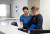 Two uniformed staff members stand behind a reception desk, smiling as they review documents in a bright, professional hospital setting.