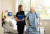 A nurse with a blue folder offers a warm smile to an elderly man with a walker in a bright, well-appointed hospital room with flowers.