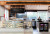A clean café with a display of pastries and baked goods, attended by a staff member. A sign reads Three Sons Espresso. Shelves and a fridge are present.