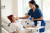 A nurse in blue scrubs checks the temperature of an older patient in a bright, comfortable hospital room with medical equipment nearby.