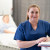 A nurse in blue scrubs holds a clipboard and smiles in a well-equipped hospital room, with a patient resting comfortably in the background.