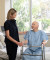 A nurse stands next to an elderly man with a walker, both smiling in a bright, welcoming hospital room with greenery outside the window.