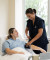 A nurse in a navy uniform checks a patient’s blood pressure in a bright, comfortable hospital room. The patient appears at ease.