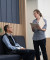 A professional woman with folders speaks to a seated man in a comfortable waiting area with wood paneling and blue seats.