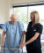 A senior man in a hospital gown walks with a walker, supported by a caring female caregiver in a well-lit private room.