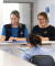 Two uniformed staff with name badges warmly assist a visitor at a reception desk, with papers and a Welcome! sign on the counter.