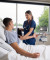 A nurse in blue scrubs calmly checks a male patient’s blood pressure in a bright, well-equipped hospital room with a view of trees.
