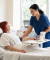 A nurse in blue scrubs gently assists an older woman resting in a hospital bed, with modern medical equipment visible nearby.