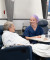 A nurse in blue scrubs and a purple cap speaks warmly with an older woman resting comfortably under a blanket in a private clinic.