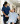 A nurse in blue scrubs holds hands with a wheelchair patient, who has a bandaged leg, in a bright, welcoming hospital corridor.