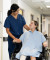A nurse in blue scrubs holds hands with a wheelchair patient, who has a bandaged leg, in a bright, welcoming hospital corridor.