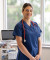 A professional healthcare worker in blue scrubs with a red stethoscope stands confidently in a well-equipped, modern medical room.