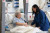 An older woman in a hospital bed smiles and holds hands with a supportive nurse in blue scrubs in a bright, modern hospital room.