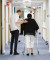 A man assists an older woman using a walker as they walk together down a well-lit hallway in a private hospital.