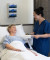A nurse in blue scrubs stands bedside, smiling and holding a clipboard, conversing with an elderly woman resting comfortably in bed.