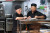 Two chefs in black uniforms calmly review notes together in a clean commercial kitchen with organized equipment in the background.