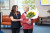 In a modern room with orange seating and large windows, two women stand together; one is supported and holds sunflowers as they smile.