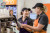 Two staff members in uniforms and caps prepare beverages together behind a coffee machine, with cups and equipment on the counter.