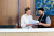 Two staff members at a reception desk review documents together, dressed professionally, with artwork displayed on the wooden wall behind.