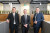 Three men in business attire stand smiling in a bright office hallway with wood accents and green plants, creating a welcoming atmosphere.