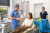 A dentist in blue scrubs consults with a smiling patient in a dental chair, assisted by staff, in a well-equipped, bright office.