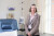 A smiling older woman in a plaid blazer stands by a blue medical chair in a well-lit hospital room with modern medical equipment.