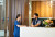 A woman in blue scrubs warmly greets a man at a white reception desk in a modern hospital with wood accents and fresh flowers.