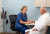A nurse in blue scrubs warmly engages an older man in a white robe during a consultation, noting details on a clipboard.