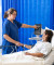 A nurse in blue scrubs attentively monitors a female patient's vital signs at her bedside in a private hospital room with blue curtains.
