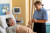 A nurse in a blue checked shirt gently checks the wrist of an older male patient resting comfortably in a hospital bed with monitoring equipment.