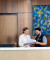 Two friendly staff members stand at a modern reception desk reviewing documents. Warm wood paneling and calming artwork are visible behind.