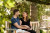 A man and a pregnant woman sit on a wooden bench outdoors, smiling with his hand gently resting on her baby bump, surrounded by greenery.