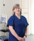 A professional, gray-haired woman in blue scrubs smiles warmly in a well-equipped, modern clinic room with medical equipment.