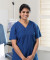 A friendly healthcare professional in blue scrubs smiles in a clean clinic room equipped with medical devices and a patient chair.