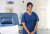 A healthcare professional in blue scrubs stands in a well-lit hospital room beside a treatment chair, IV equipment, and window blinds.