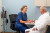 A nurse in blue scrubs chats with an older man in a white robe on an exam table in a modern, well-equipped medical office.