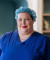 A healthcare professional in blue medical scrubs and a surgical cap stands indoors, smiling gently in a calm, modern clinical setting.