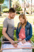 A man and woman stand together outdoors, smiling at their newborn in a hospital bassinet. The baby is safe in a striped blanket.